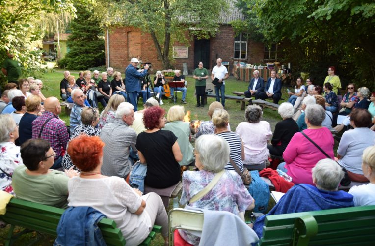 Grupa osób w starszym wieku siedzących w półokręgu (tyłem do obiektywu) na świeżym powietrzu. Frontem stoją dwie osoby, mężczyzna i kobieta. Na lewo od nich mężczyzna z kamerą. Za plecami mają parterowy budynek z cegły. Na drzewach liście są zielone.